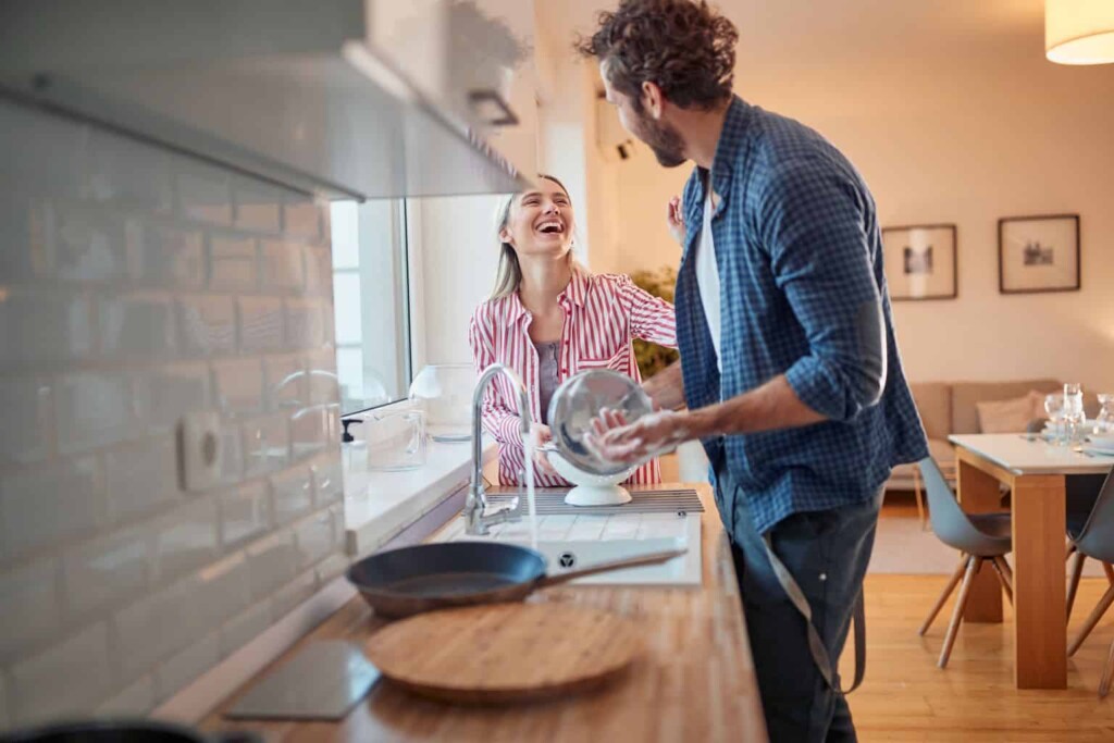 A couple laughing while washing dishes in a brightly lit kitchen